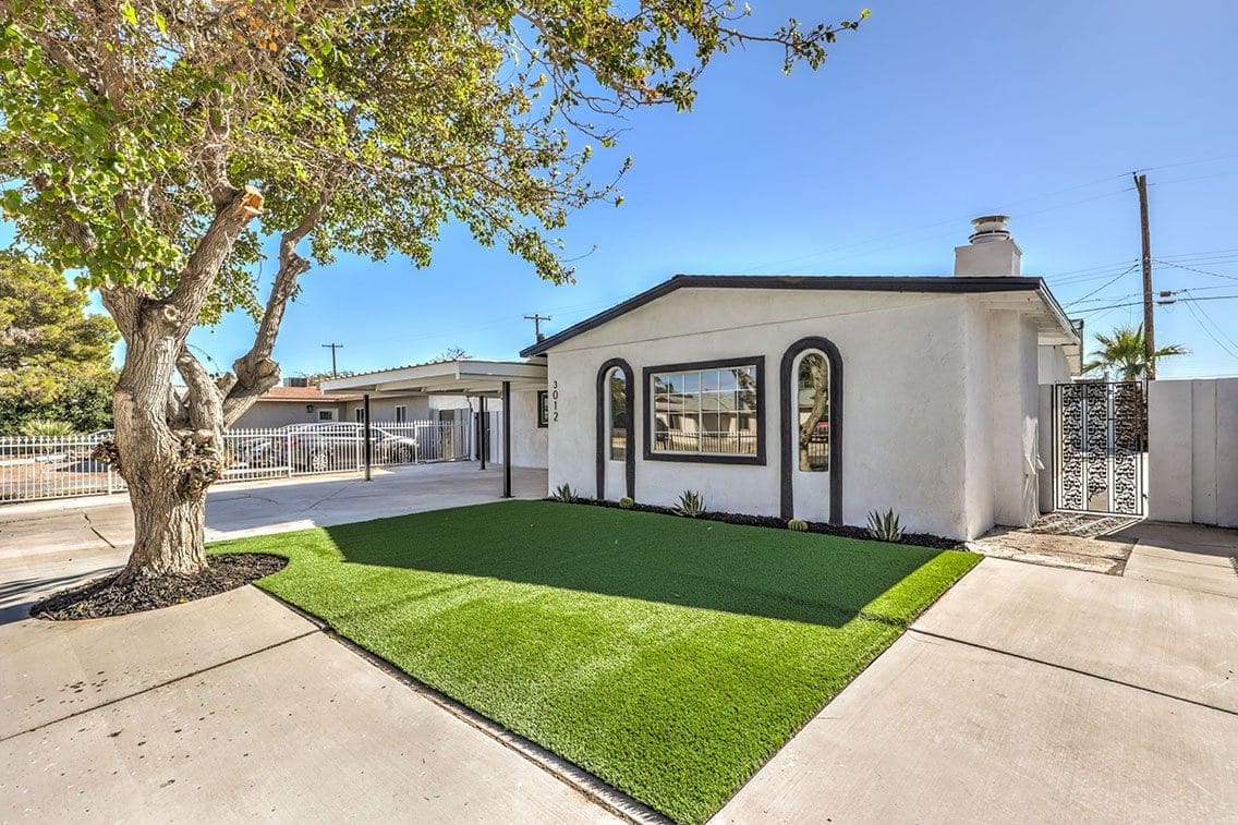 A single-story white house with black trim, a large window, an artificial grass lawn, a tree on the left, and a gated driveway under a clear blue sky.