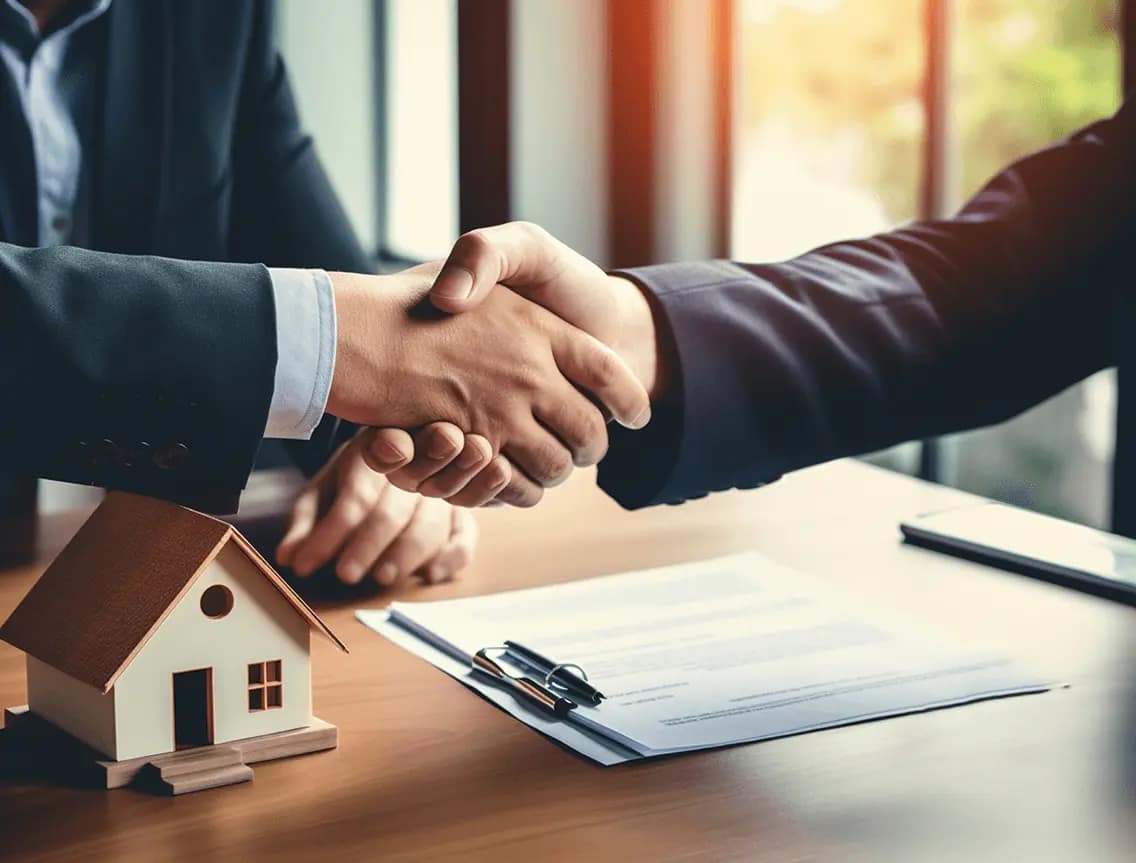 Two people in business suits shake hands across a desk with a small model house, a pen, and documents, suggesting a cash buyer finalizing a real estate agreement to help you sell your house fast.