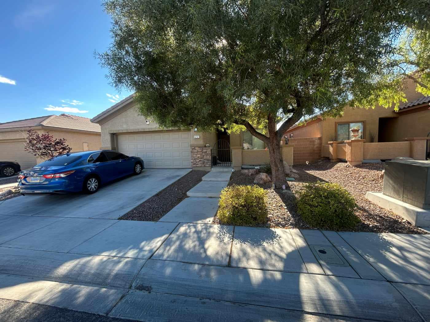 A suburban house with a two-car garage, a blue sedan in the driveway, and desert landscaping—with Better Home Buyer, you can sell my home as is under the clear sky and lush tree in the front yard.