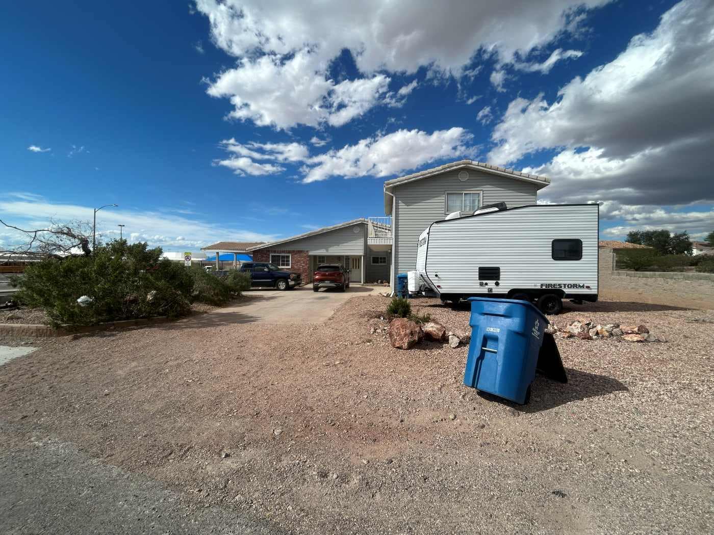 A house with a gravel driveway, a blue recycling bin, an RV trailer, several parked vehicles, desert landscaping, and a partly cloudy sky above—perfect if you want to sell my home as is to a Better Home Buyer.