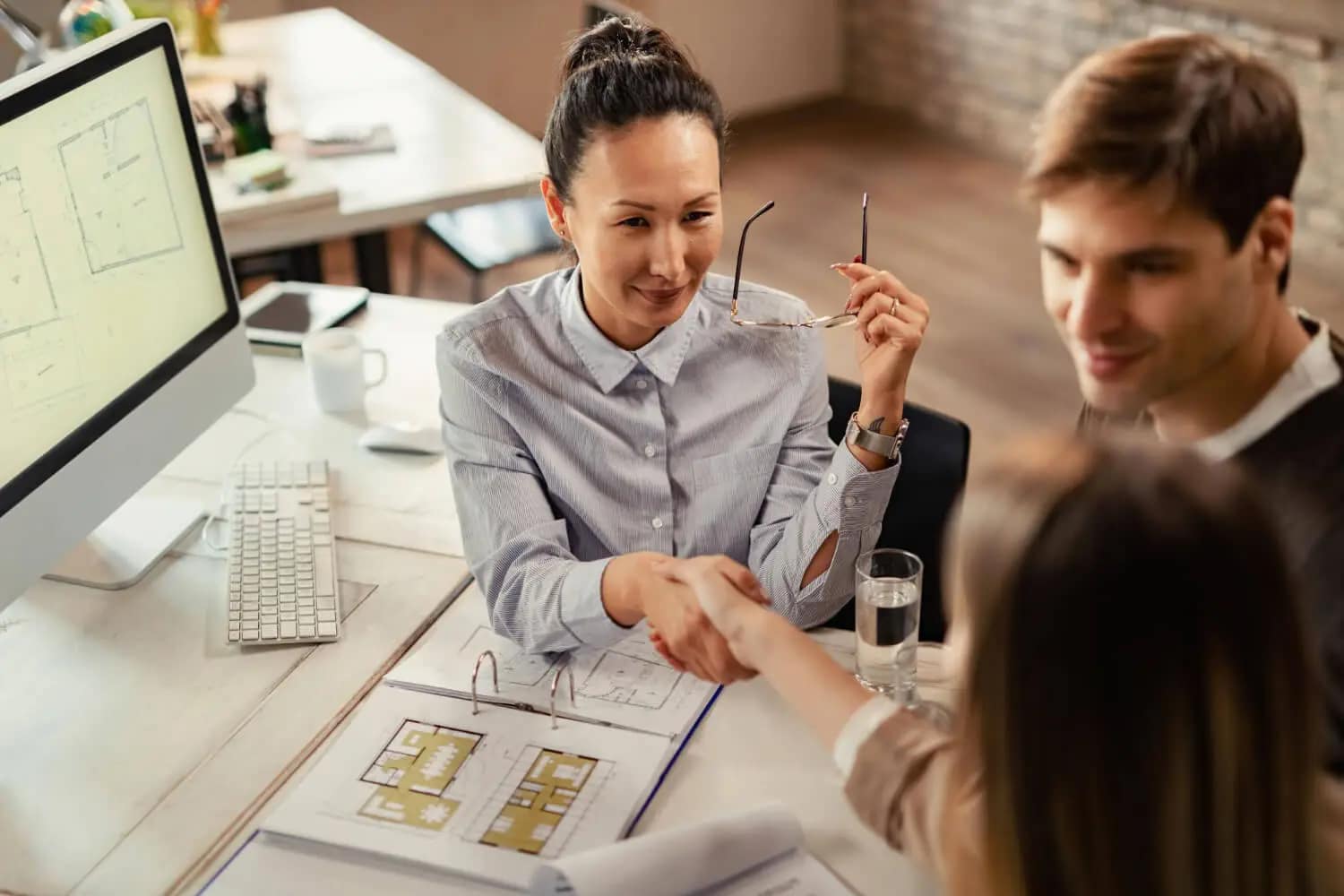 A woman in business attire shakes hands with another woman across a desk, smiling. A man sits beside them. Architectural plans are visible, suggesting a meeting where a cash buyer is ready to buy houses just as they are.