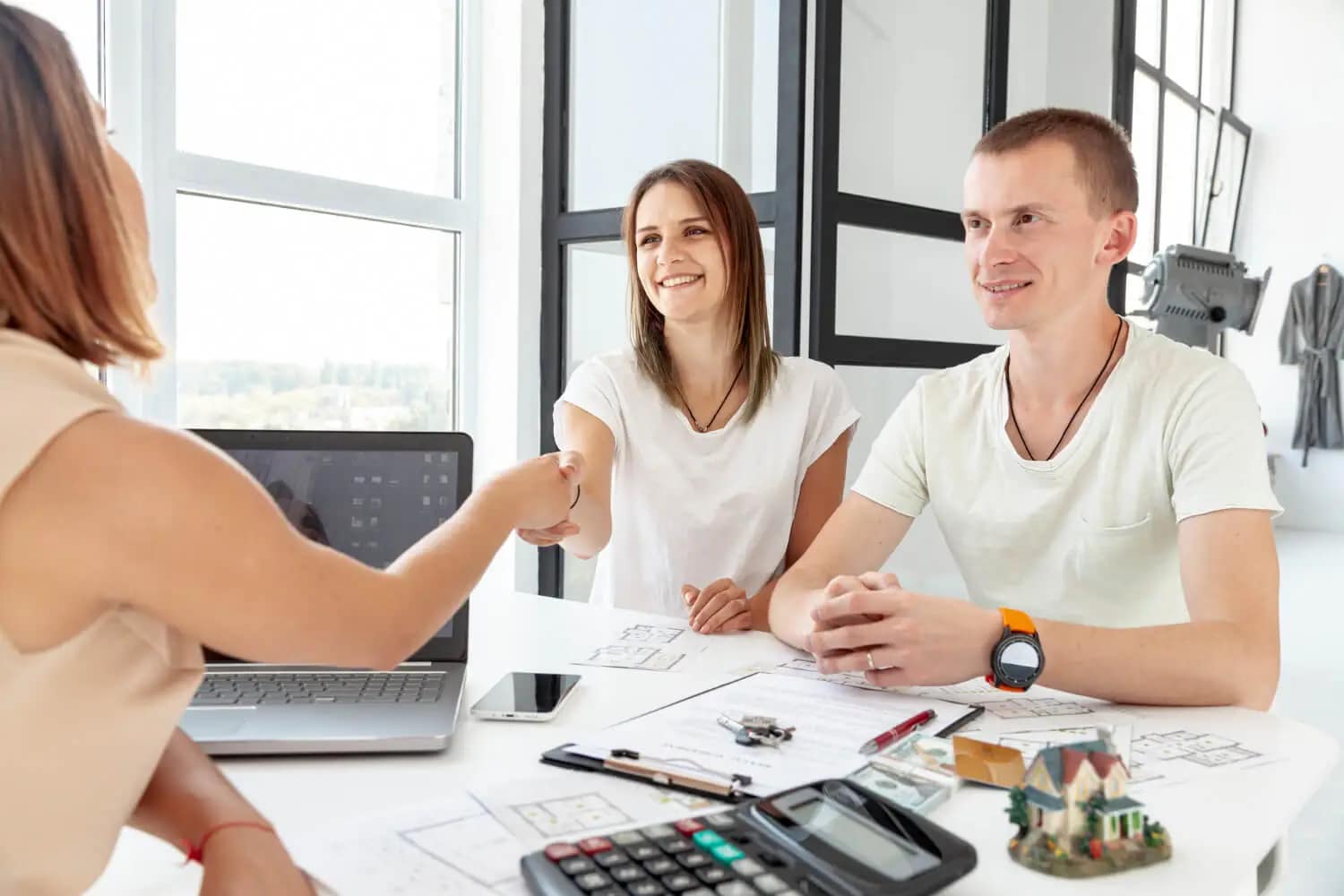 A woman shakes hands with another woman across a desk while a man smiles beside them. Papers, a laptop, a calculator, and a model house hint at a real estate meeting—perfect for those looking to sell your house fast or sell your house as is.