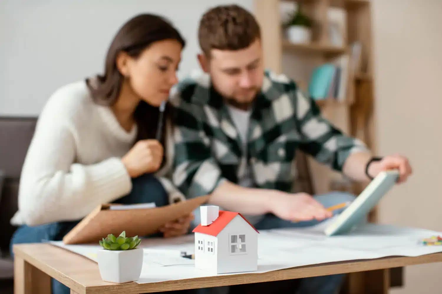 A woman and man sit at a table looking at papers and a tablet, appearing focused. A small model house and a potted plant are on the table in the foreground—perhaps they're finding ways to sell your house fast in their living room.