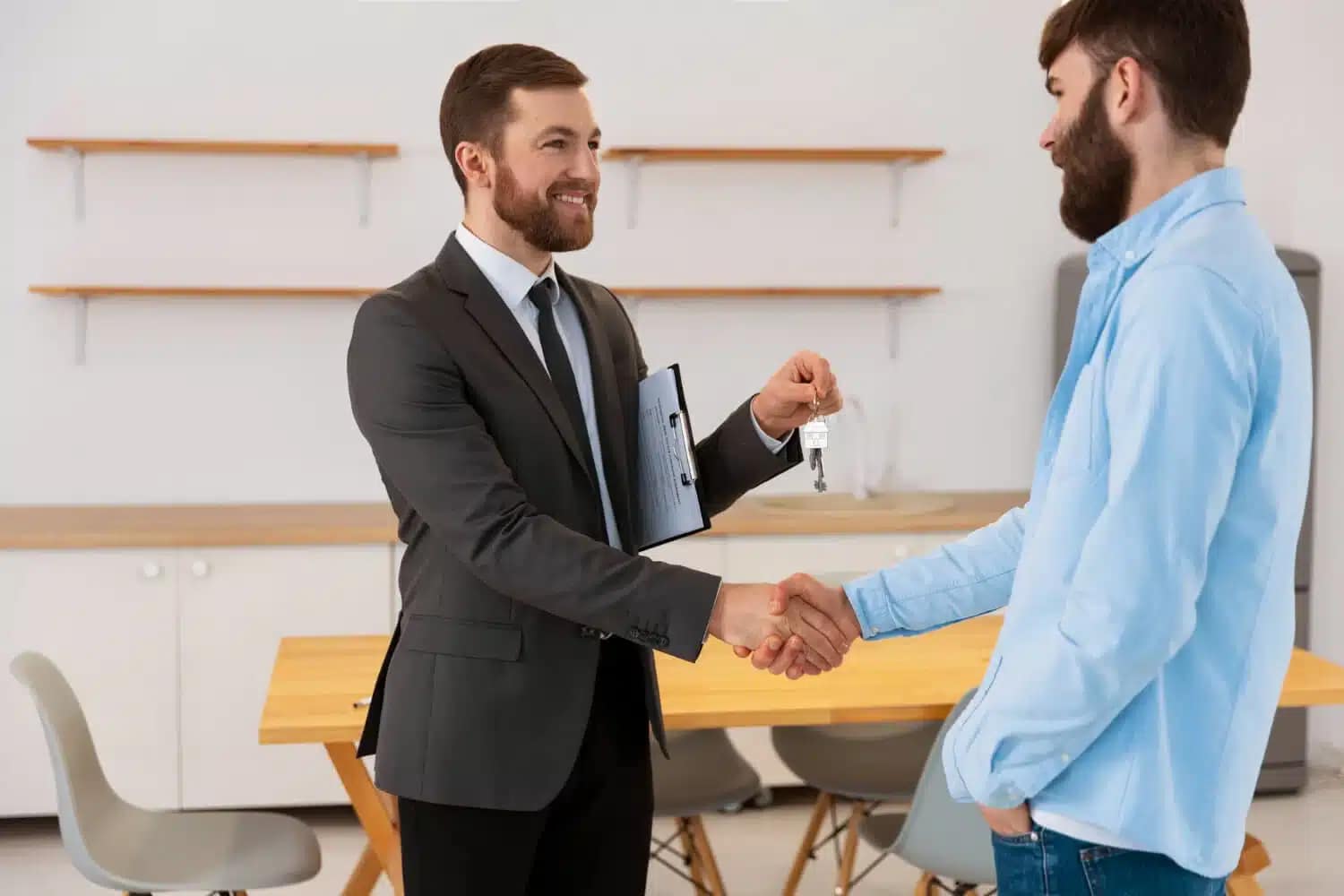 A cash buyer in a suit hands over a set of keys to another man in casual clothing while they shake hands in a modern, bright room with a table and chairs in the background.