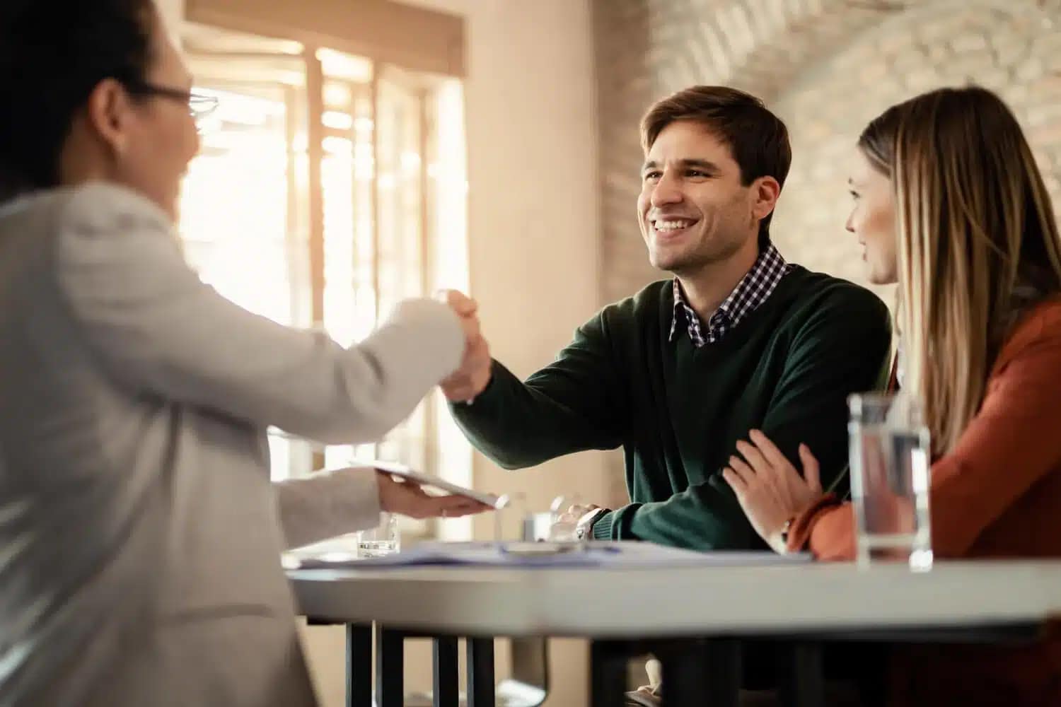 A smiling man, representing a cash buyer, shakes hands with a woman across a table while another woman looks on and smiles. Papers and a glass of water rest on the table in this well-lit room.