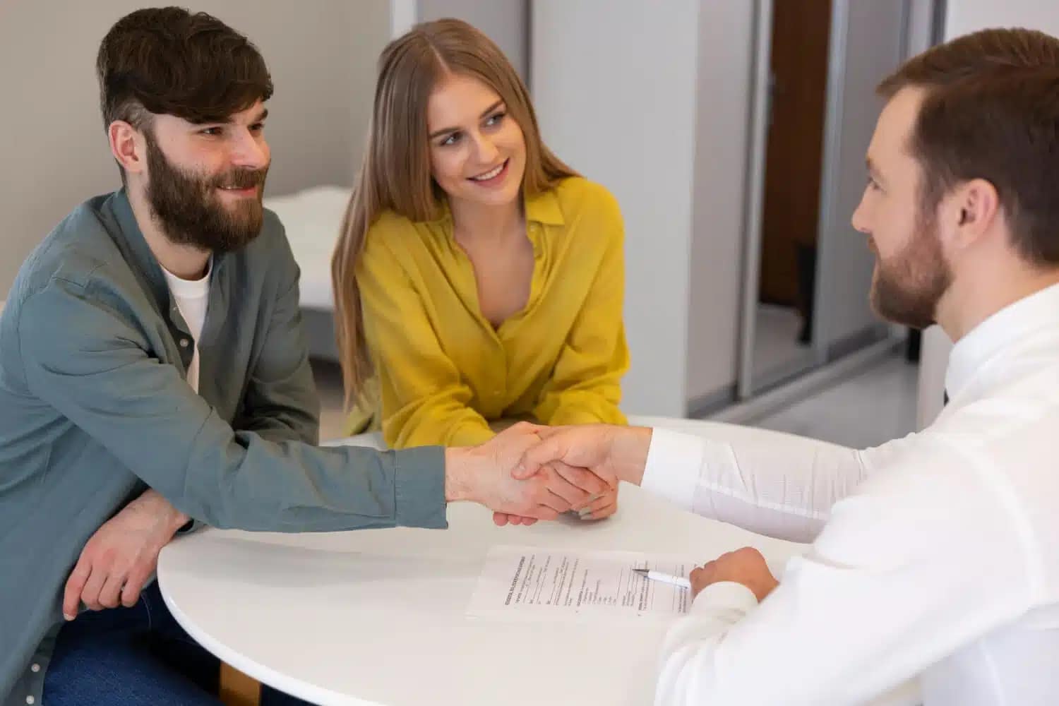A man and woman sit at a table, smiling, while the man shakes hands with a cash buyer in a white shirt across the table. A document with text and a pen lies on the table in front of them.