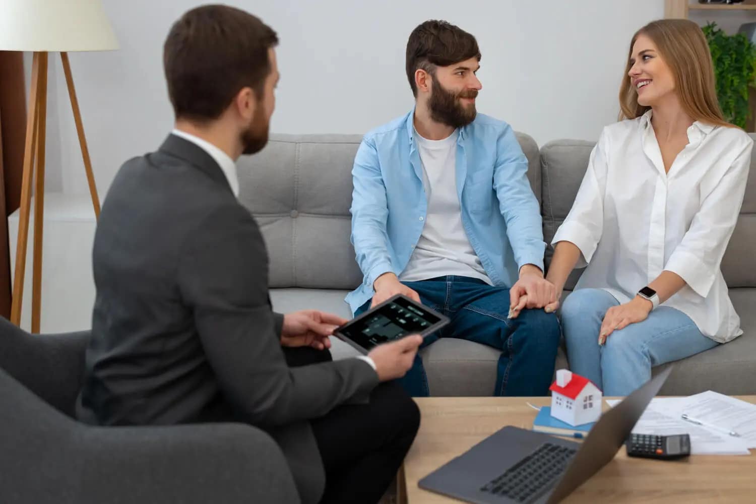 A man in a suit holding a tablet sits across from a smiling couple on a couch. A small model house and paperwork on the coffee table suggest they're discussing how to sell your house fast in a real estate or financial meeting.