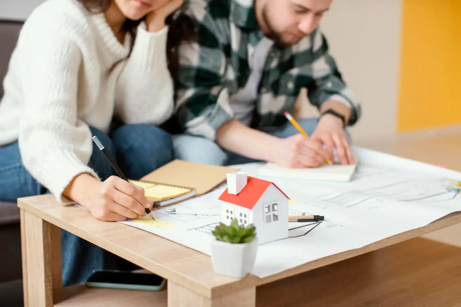 A woman and a man sit at a table, writing and drawing on papers with a small model house and a potted plant in the foreground—perhaps planning how to sell your house fast or work with a cash buyer for their next home project.