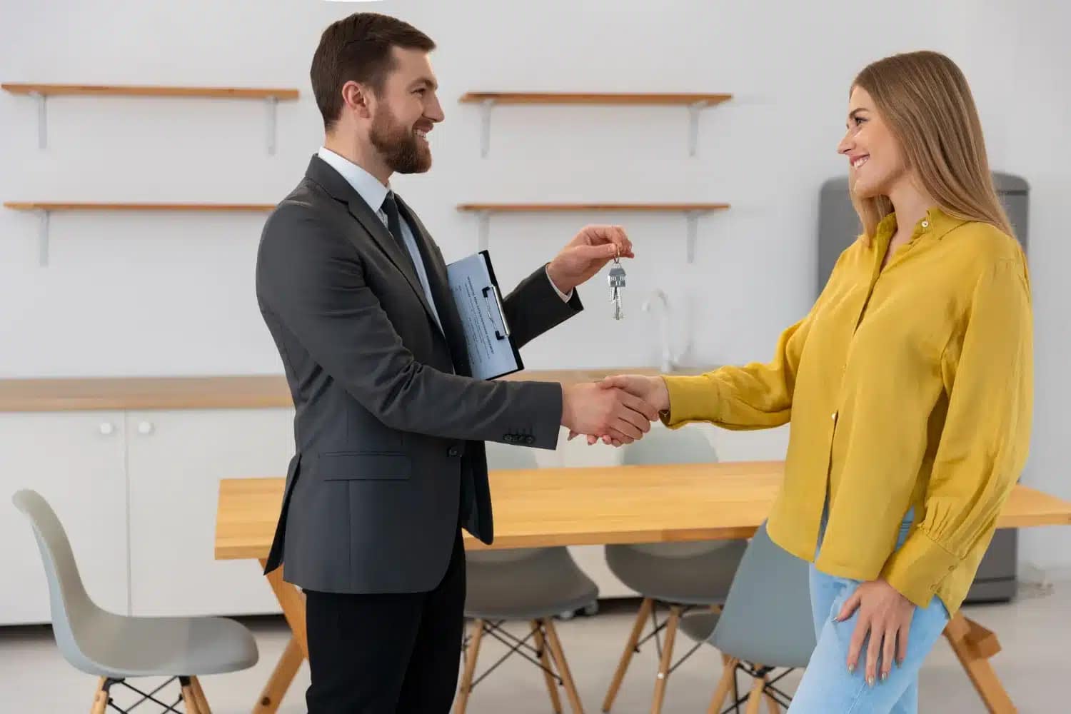 A cash buyer in a suit hands keys to a smiling woman while they shake hands in a modern, minimalist room with a wooden table and chairs.