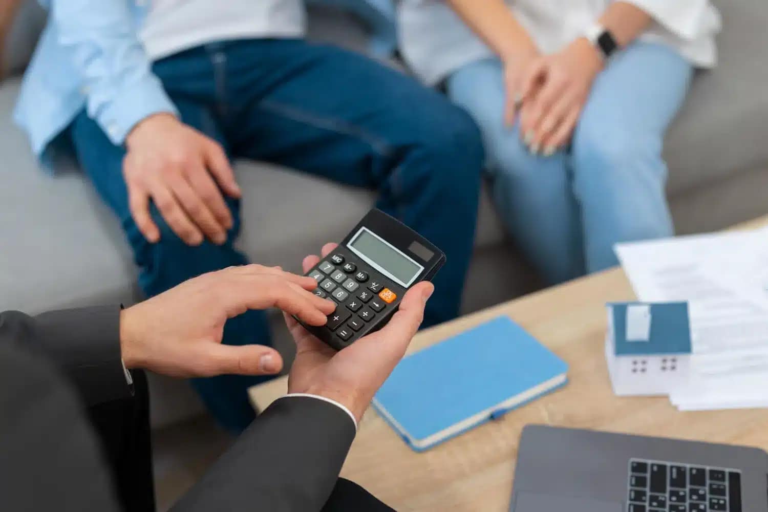 A person in a suit uses a calculator at a table while two casually dressed people sit nearby; documents, a notebook, and a laptop suggest a financial meeting—perhaps discussing how to sell your house fast to a cash buyer.