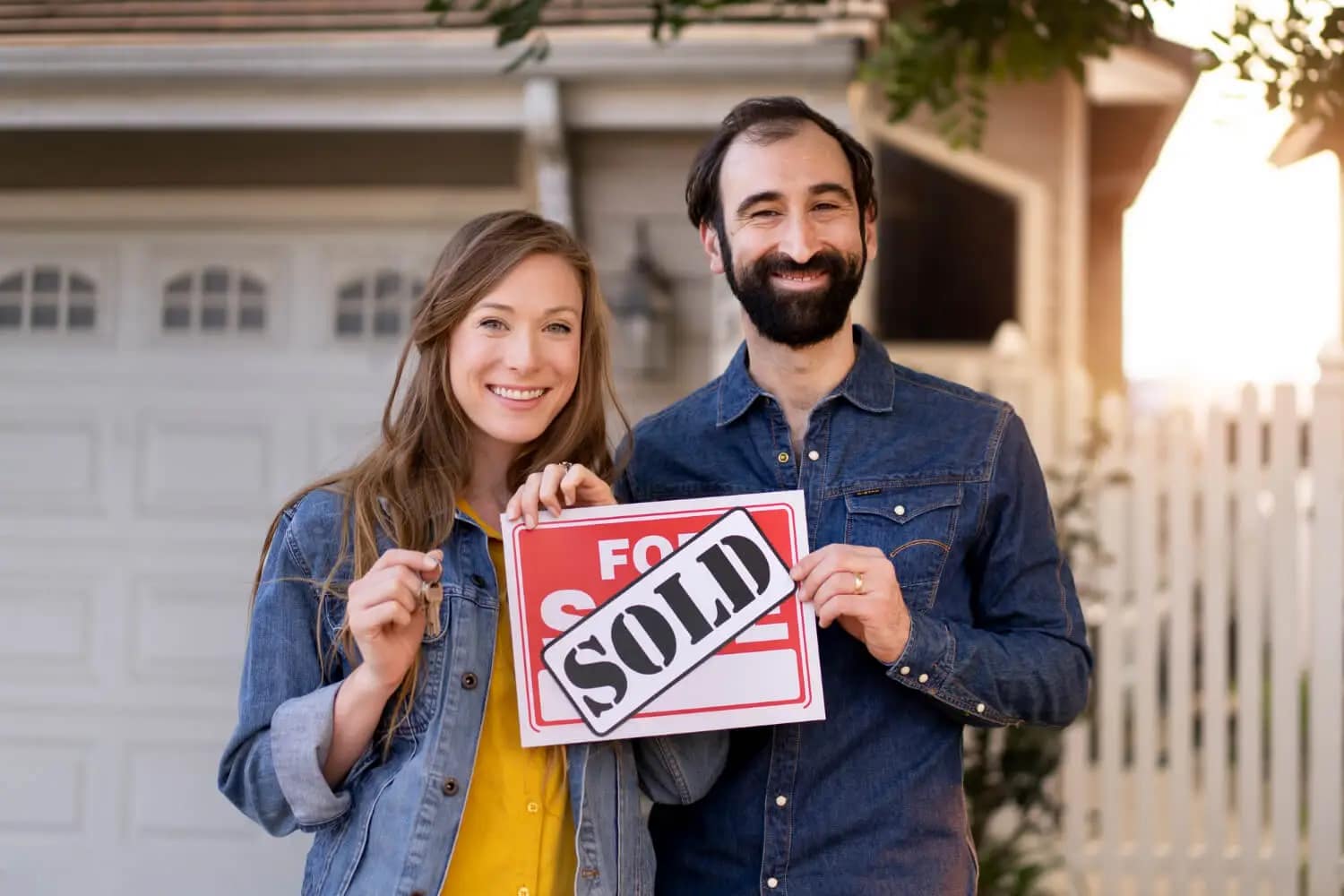 A smiling couple stands in front of a house, holding a For Sale sign with a SOLD sticker—proof that you can sell your house fast. They look happy in casual clothes, with the house and garage door in the background.
