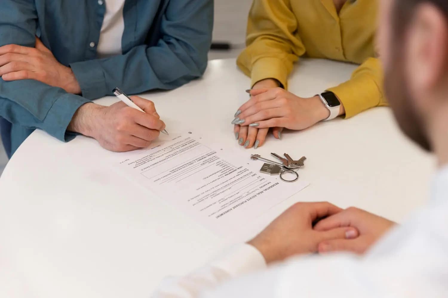 A person signs a document on a table while another sits beside them. Keys rest on the table, suggesting a real estate or rental agreement—perhaps with a "We buy houses" service to help you sell your house fast. Another individual is partially visible across the table.