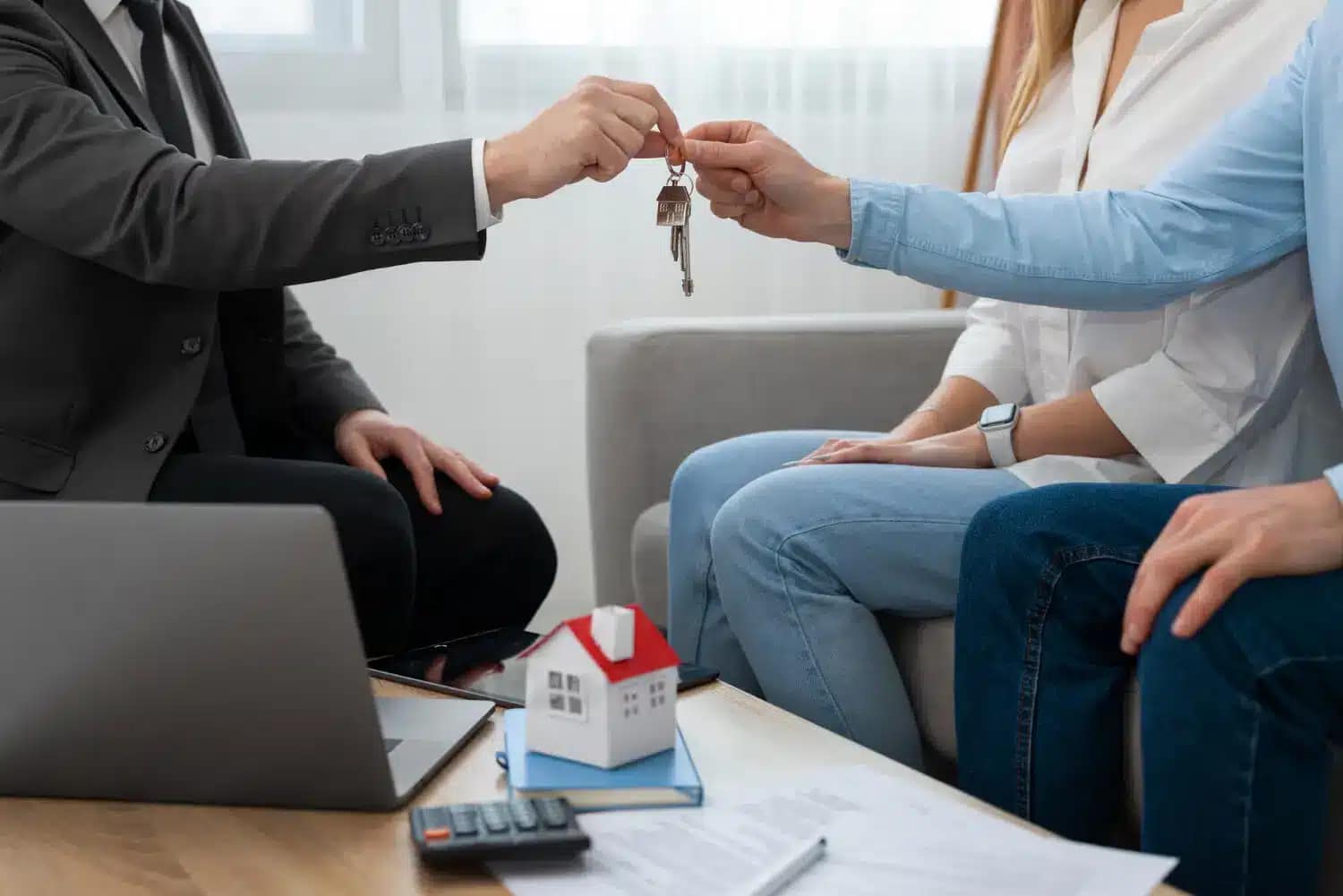 A person in a suit hands house keys to a couple sitting on a couch, as they review documents and a house model—illustrating a real estate transaction where you can sell your house as is to a cash buyer.