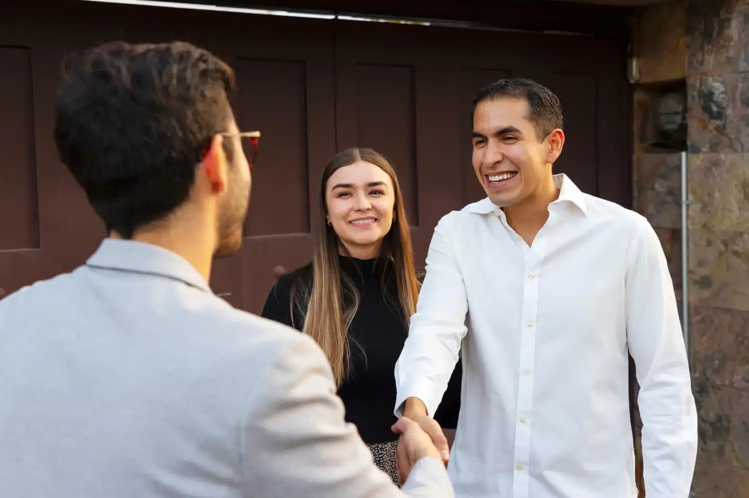 A man in a white shirt smiles while shaking hands with another man in a light jacket. A woman with long brown hair stands beside them, also smiling—celebrating a smooth "We buy houses" deal outside a wooden gate and stone wall.