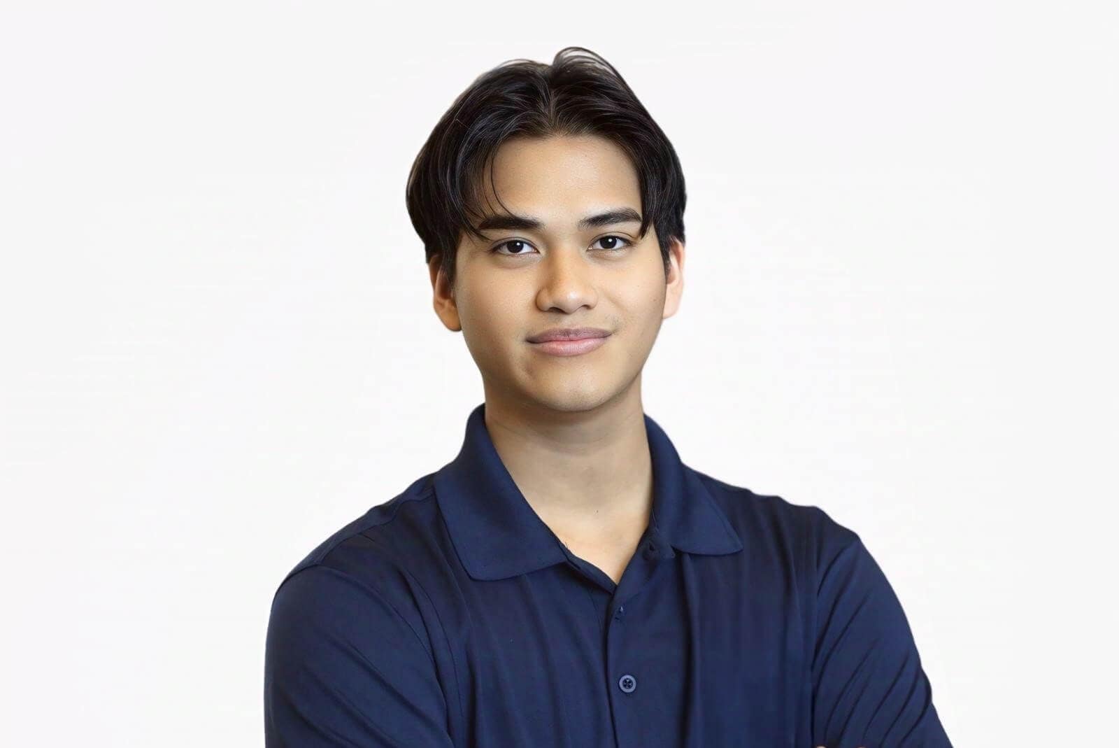 A young man with medium-length dark hair wearing a navy blue collared shirt stands against a plain white background, looking at the camera with a slight smile and arms crossed—embodying confidence like Better Home Buyer clients.