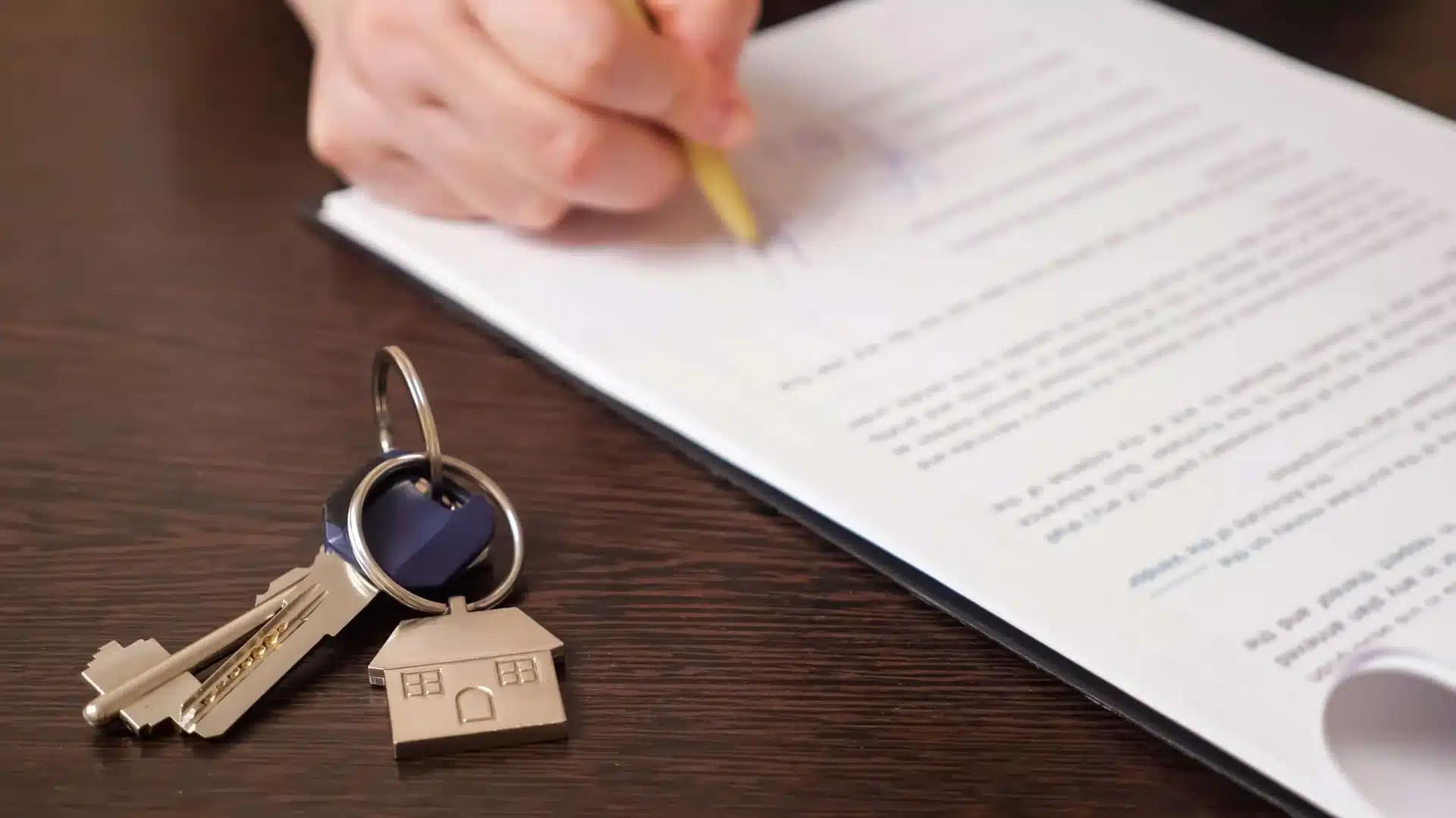A person signs a document with a pencil on a wooden table; nearby, a keyring with a house-shaped keychain and two keys rests—an ideal scene for cash buyers ready to help you sell your house fast.