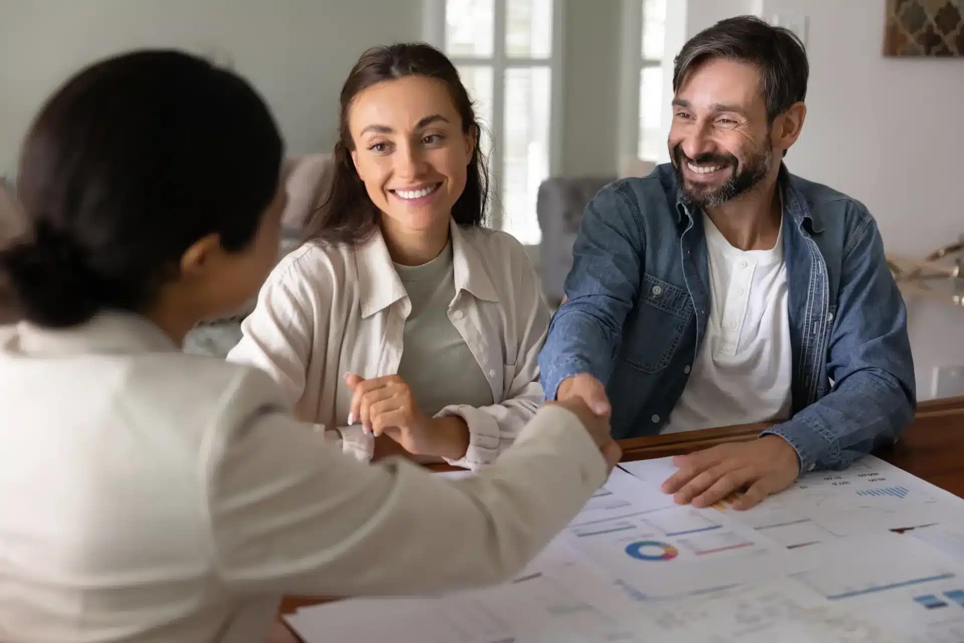 A professional shakes hands with a smiling man while a woman beside him also smiles. They are sitting at a table covered with documents and charts, suggesting a successful "sell your house as is" business meeting or agreement.