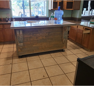 A kitchen with wooden cabinets, a tiled floor, and a marble-topped island. On the island are a large water jug and a bottle. Sunlight shines through windows above the sink.