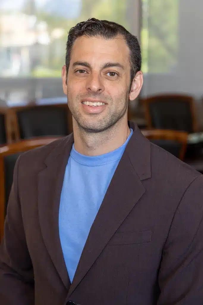 A man with short dark hair wearing a brown blazer over a light blue shirt smiles at the camera in a bright office, ready to tell you more About Better Home Buyer and how they help create better home buyers.
