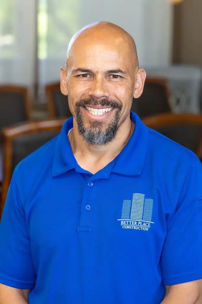 A smiling man with a bald head, goatee, and mustache wears a royal blue Better Place Construction polo. He sits indoors—blurred chairs and windows behind him—ready to share more about Better Home Buyer experiences.