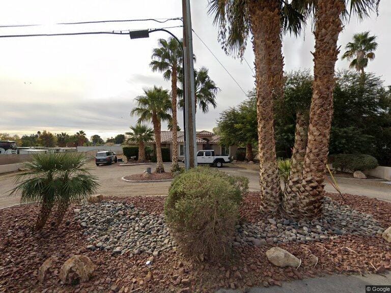 A suburban house with a tile roof and a white SUV in the driveway, surrounded by palm trees, rocks, and desert landscaping under a cloudy sky—perfect for those looking to sell my home as is to Better Home Buyer.