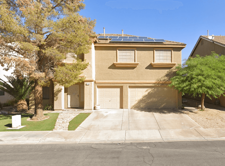A beige two-story suburban house with solar panels on the roof, two garage doors, and a driveway, surrounded by trees and a desert landscape under a clear blue sky.