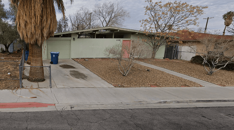 A small, single-story house with a green exterior, red door, and attached garage. The front yard has gravel, a few leafless trees, and a blue recycling bin by the driveway.