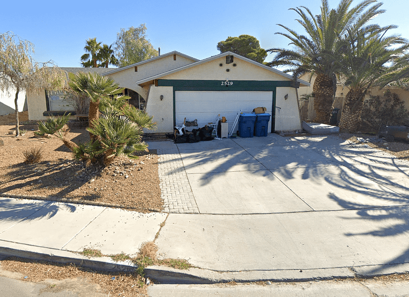 A single-story house with a green and white garage door, trash bins, and bags on the driveway. Palm trees and desert landscaping surround the front yard under a clear sky.