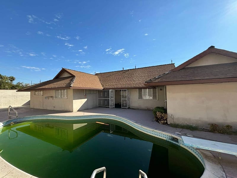 A single-story house with a brown roof and barred windows sits behind a backyard pool filled with green water. The sky is clear and blue.