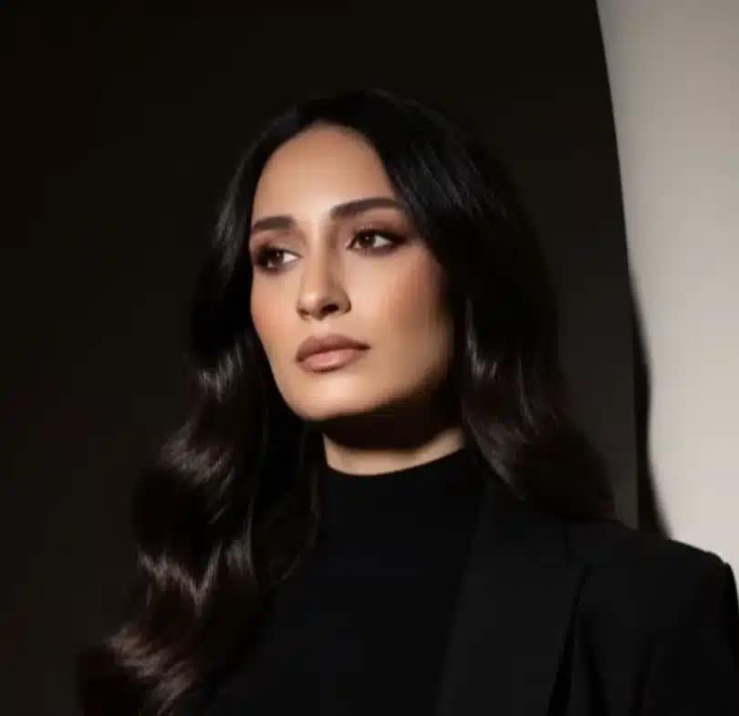 A woman with long, wavy dark hair stands against a curved, light-colored wall, wearing a black blazer and matching turtleneck. She looks to the side with a serious expression, as if considering better home buyers and their impact.