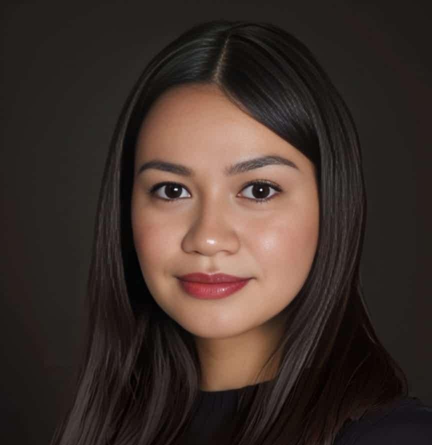 A young woman with long, straight dark hair and clear skin poses against a dark background, wearing a dark top and a soft smile on her face.